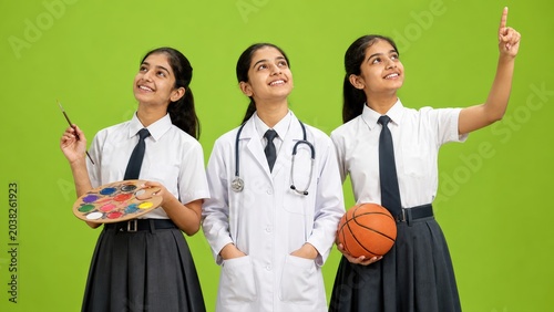 Three smiling Indian schoolgirls dream about future careers while holding symbols for art medicine and sports against a bright green background