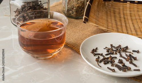 A small glass containing clove tea, and a few cloves in white container on the table.