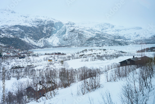 Beautiful winter landscape in Norway featuring traditional wooden houses and cabins nestled in a snow-covered valley with majestic mountains and a frozen lake under a bright Arctic sky.