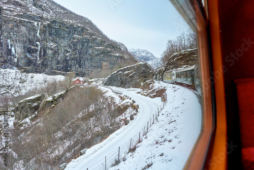 Scenic winter railway journey through the snow-covered fjords and mountains of Norway, viewed through a train window showing the curved track, a traditional red house, and rugged cliffs.