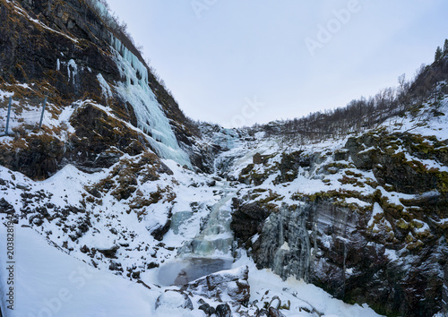 Breathtaking frozen waterfall and ice formations on a steep rocky mountain cliff during winter in Norway. Scenic arctic landscape with snow-covered terrain viewed from a famous scenic train route.