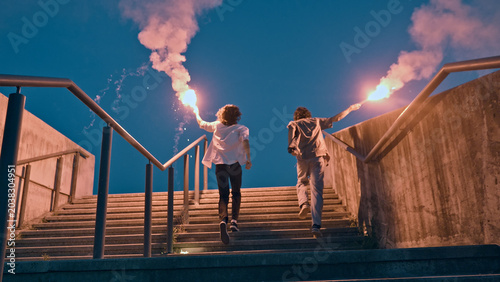 Foto Two skateboarders, silhouetted against the twilight sky, descend a concrete staircase holding flaming torches, their movements carefree and rebellious