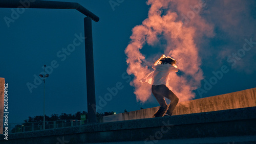 Photos A skateboarder, engulfed in adolescent rebellion, lights a flare, its radiant glow illuminating his silhouette against the twilight sky, symbolizing the carefree, defiant essence of youth culture