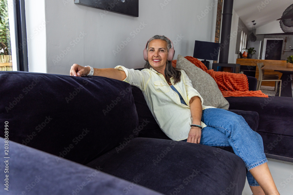 Fototapeta premium Senior woman relaxing sideways on dark velvet sectional at home, wearing pink headphones, smiling