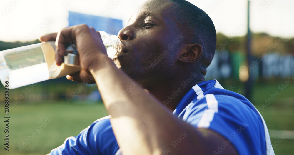 Naklejka premium Soccer, break and black man with drinking water for hydration, wellness and training outdoor. Football, thirsty and sports person or player with liquid bottle, resting and practice on field for game
