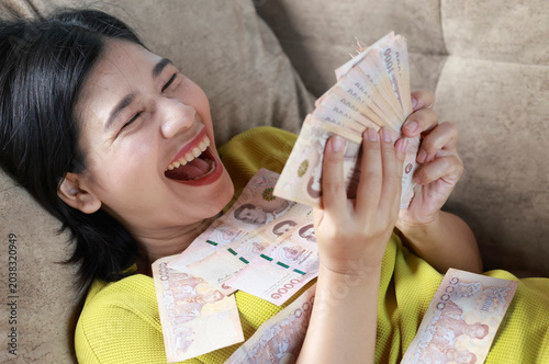 Happy Asian woman counting cash lying on sofa, Thai baht banknote, money, cash, financial success, income, savings, bonus, wealth, and a comfortable lifestyle, close-up