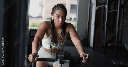 Athletic woman with braided hair and tattoos intensely working out on a stationary exercise bike, building stamina and endurance during a cardiovascular training session in a modern fitness center