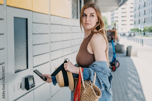 Woman holds smartphone near automated package terminal and shopping bags. Smart city retail infrastructure, urban technology integration service, automated neighborhood locker hub.