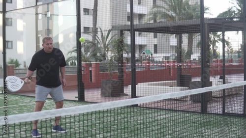 Man playing padel on an outdoor court in a residential area, preparing to hit a low ball near the net. Casual sports lifestyle scene showing an amateur player during a friendly match or training