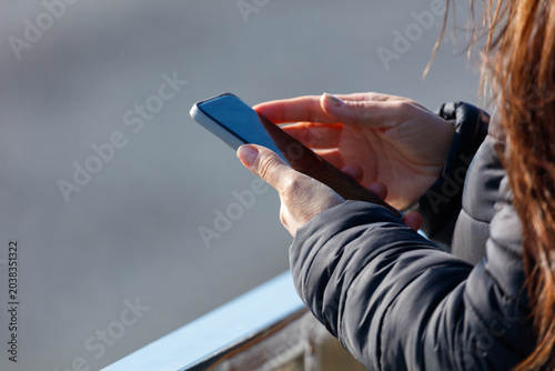 Woman holds smartphone outdoors. Fingers tap screen with focus. Dark jacket covers arms. Hair strands drift in breeze.