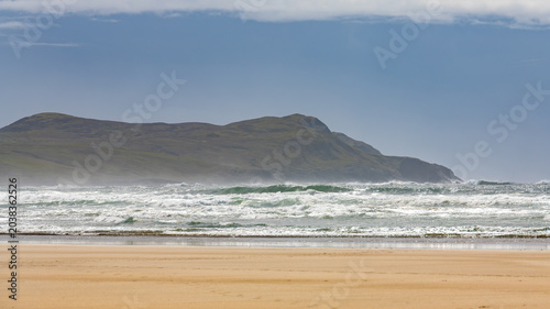 Photos Scenic view of Machir Bay Beach on the Isle of Islay in Scotland, featuring powerful ocean waves rolling onto the sandy shore with rugged cliffs in the background