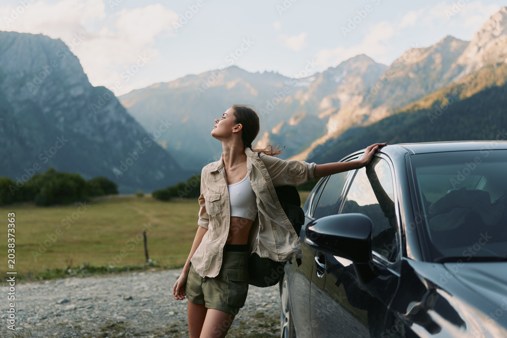 Fototapeta premium Young woman in casual clothes leans on a car and enjoys fresh mountain air with closed eyes during a peaceful outdoor moment in nature.