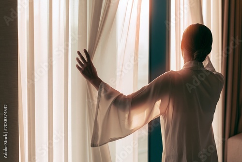 Woman in robe opening sheer curtains at sunrise