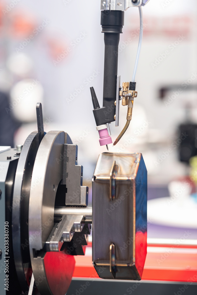 Ταπετσαρία Industrial TIG welding torch positioned over a clamped angular metal workpiece held in a precision chuck, demonstrating automated arc welding setup at an indoor manufacturing exhibition