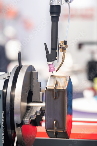 Obraz na plátně Industrial TIG welding torch positioned over a clamped angular metal workpiece held in a precision chuck, demonstrating automated arc welding setup at an indoor manufacturing exhibition