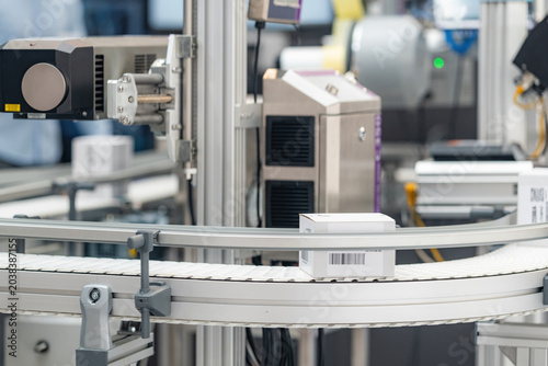 Automated industrial packaging and labeling system processes small white boxes on a curved conveyor belt, captured during a live demonstration at an indoor manufacturing technology exhibition.