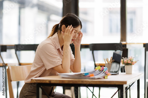 Stressed young asian business woman suffering from a severe headache or migraine while working at her office desk. Overwhelmed by work pressure. Fatigue. And burnout