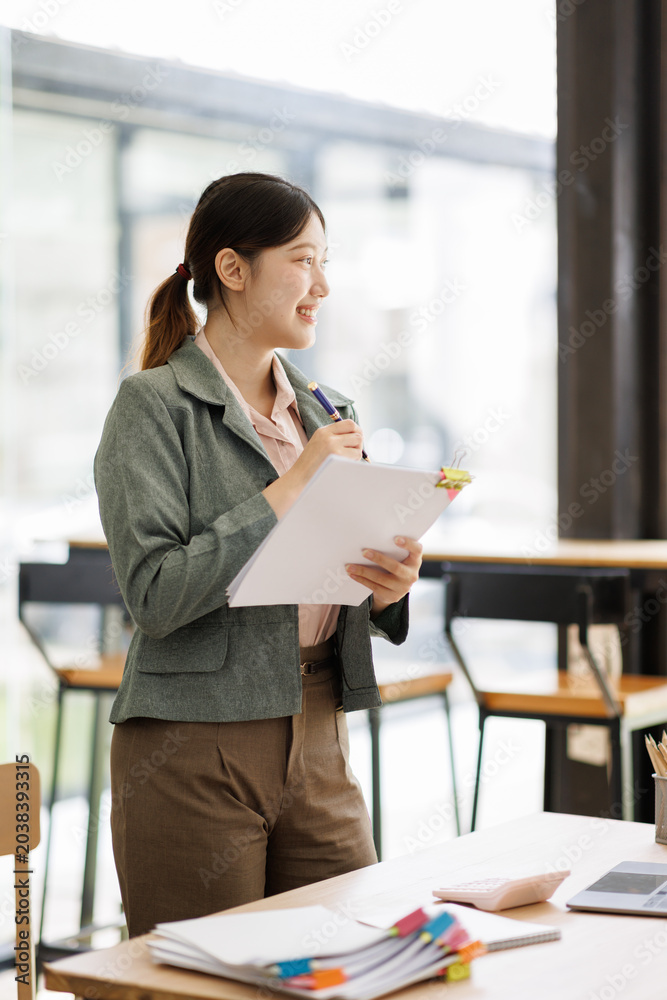 Obraz premium Asian businesswoman standing in modern office, smiling while holding papers and pen, writing and organizing documents to plan, manage tasks and drive career growth
