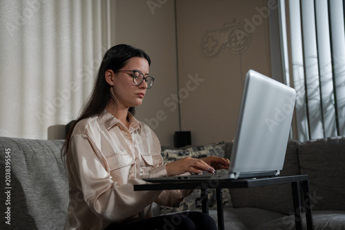 Young Woman Wearing Glasses Working On Laptop At Home sitting on a sofa and focusing on remote work tasks in a comfortable domestic environment