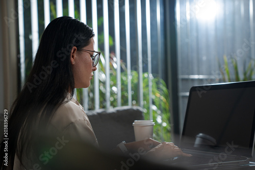 Young Woman Working On Laptop Near Window At Home with a coffee cup while focusing on remote tasks and maintaining productivity in a comfortable environment
