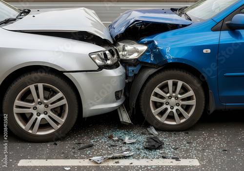 Silver and blue cars in a serious head on road collision accident on an asphalt street with broken glass debris