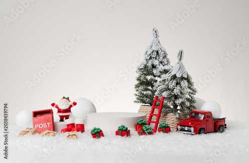 Konstfotografi Christmas product display photography featuring a white cylindrical podium, red toy truck, Santa Claus figurine, red mailbox, wooden ladder, ornament ball, and wrapped gift boxes on artificial snow