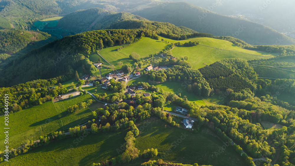 Fototapeta premium Aerial view of small rural village among green hills forests and fields during golden sunset light in German countryside