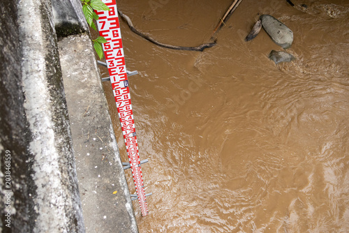Konstfotografi Red and white water level gauge measuring murky brown floodwater rising against
