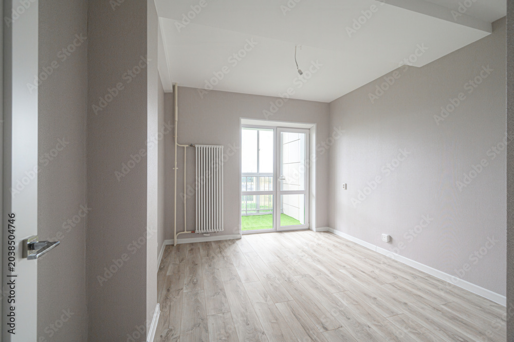 Obraz premium A bright empty room with light grey walls, light wood laminate flooring, white ceiling, a white radiator, and a large glass door leading to a balcony
