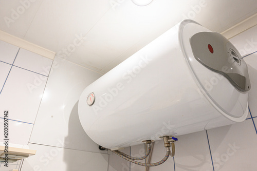 A white horizontal electric water heater mounted on a tiled wall, featuring a pressure gauge, control panel, and flexible hoses connected at the bottom