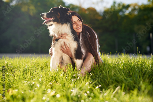 Bild auf Leinwand Woman with long dark hair sits on grass, embracing a black and white dog in a su