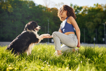 Young woman with curly hair kneeling on grass interacts with a black and white dog in a sunny outdoor park setting surrounded by trees and greenery