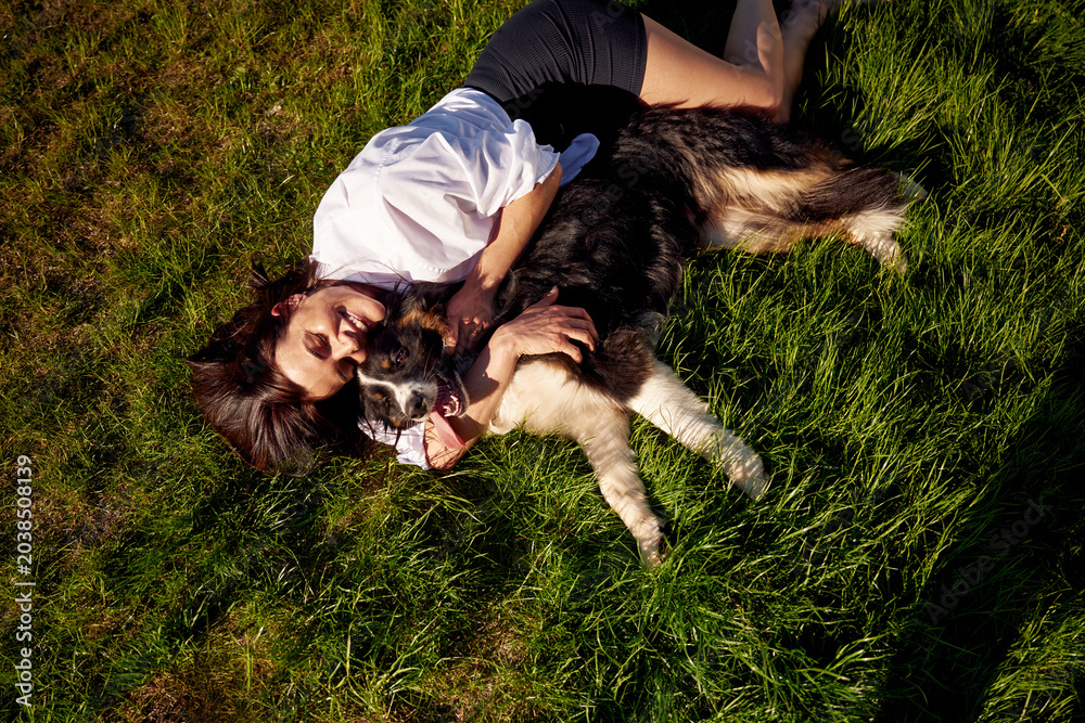 Obraz premium Woman lying on green grass with a dog, embracing and playing together in a sunny outdoor setting, showcasing a joyful bond between pet and owner