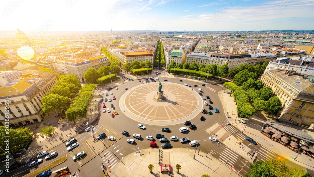 Fototapeta premium Aerial view of a bustling city street featuring a prominent roundabout with a statue at its center, surrounded by lush trees and historic buildings, showcasing urban landscape and architecture.