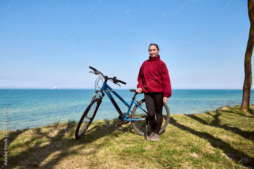 Fototapeta premium Young woman standing with blue bicycle on grassy coast under clear blue sky