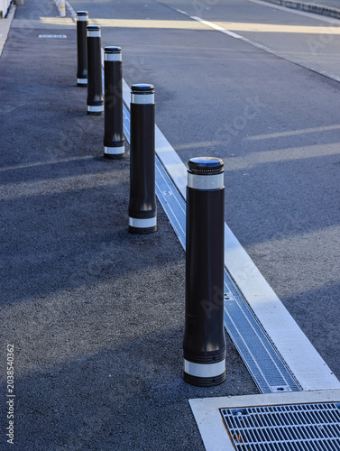 The brown metal bollards with white reflective strip installed along the roadside near a modern residential area. The bollards act as vehicle barriers for traffic control and pedestrian safety.