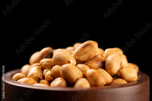 Pile of roasted hulled peanuts in brown wooden bowl on black background wiht copy space for text. Close-up