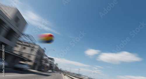 Motion blurred colorful beach ball flying fast through a bright blue sky over a city street with buildings and cars