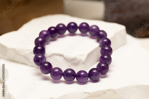 A circular bracelet made of purple amethyst stones sits on a white stone surface. The stones are smooth and round, reflecting light in a simple setting.
