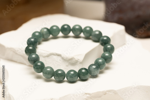 A simple bracelet made of Burmese Jade sits on a white stone surface. The beads are round and smooth. The setting highlights the natural colors of the jade in this display.