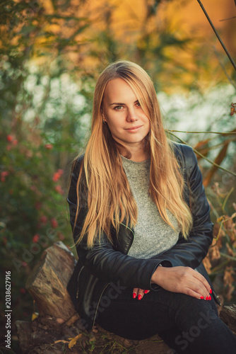 Portrait of young blonde woman in black leather jacket sitting outdoors in autumn park