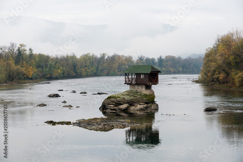 Small wooden house on rock in middle of Drina river near Bajina Basta in Serbia during cloudy morning with fog obscuring mountain in background