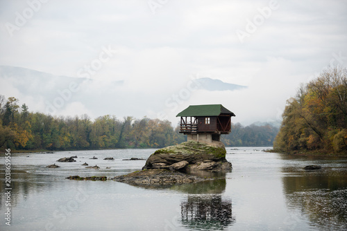 Small wooden house on rock in middle of Drina river near Bajina Basta in Serbia during cloudy morning with fog obscuring mountain in background