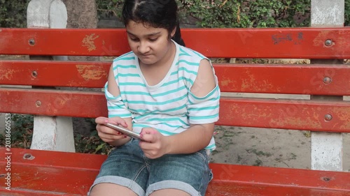  Young Indian girl sitting on a wooden bench and using a smartphone outdoors during summer time in India. Concept of mobile addiction, digital entertainment, screen time, and modern childh