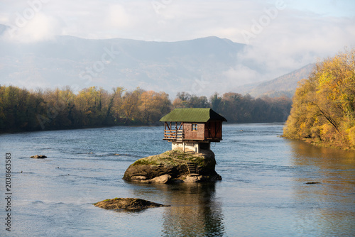 Small wooden house on rock in middle of Drina river near Bajina Basta in Serbia during sunny day with some fog leftovers before mountain in background
