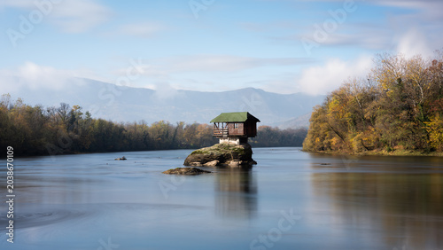 Small wooden house on rock in middle of Drina river near Bajina Basta in Serbia during sunny day with some fog leftovers before mountain in background