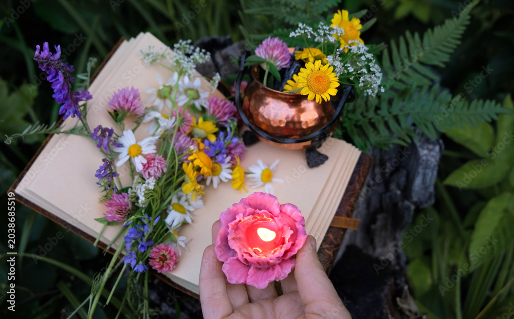 Obraz premium hand with burn floral shape candle, magic book, copper pot and wild flowers in forest, abstract nature background. Green witch, practical magic, esoteric ritual, witchcraft. close up