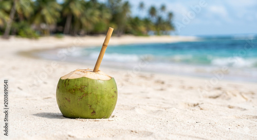 Billede på lærred Fresh green coconut drink with a straw on a tropical sandy beach with turquoise sea and sky background