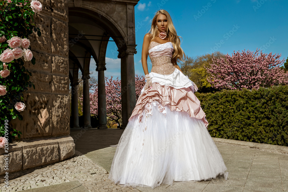 Fototapeta premium Stunning blonde fashion model in an extravagant white and pink ballgown posing outdoors under a bright blue sky with blooming spring blossoms and stone arches.