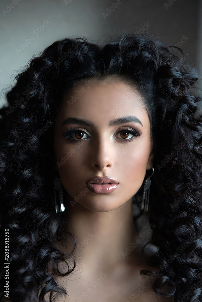 Fototapeta premium A beautiful woman with long, dark, curly hair is posing for a portrait against a plain background.